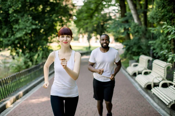 Casal multiétnico atlético, homem de pele escura e mulher de cabelos vermelhos caucasianos, correndo juntos. Os corredores do esporte jogging na trilha do parque na manhã adiantada. Foco na mulher — Fotografia de Stock