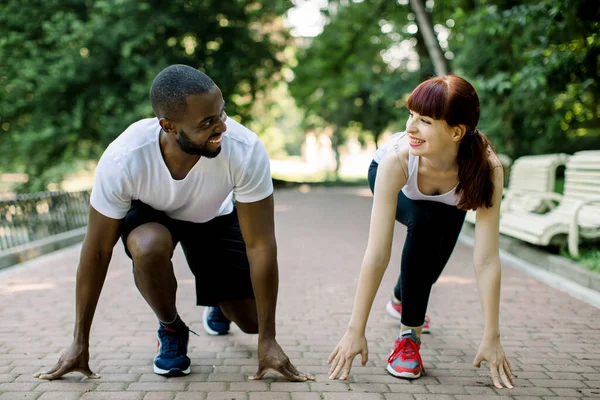 Multiethnic couple in sport clothes on starting line preparing to run in the park, smiling and looking each other. Healthy fit and sportive couple starting position ready for running