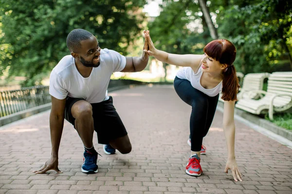 Casal fitness jovem de homem barbudo Africano e mulher caucasiana atraente, pronto para começar a correr no parque, dando alta cinco uns aos outros e sorrindo. Jogging juntos ao ar livre — Fotografia de Stock
