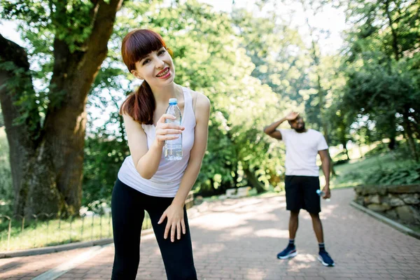 Närbild av vackra kaukasiska kvinnliga löparen ler mot kameran och håller vattenflaska, vilar efter hård träning i parken. Trött afrikansk man på bakgrunden, har rast efter jogging — Stockfoto