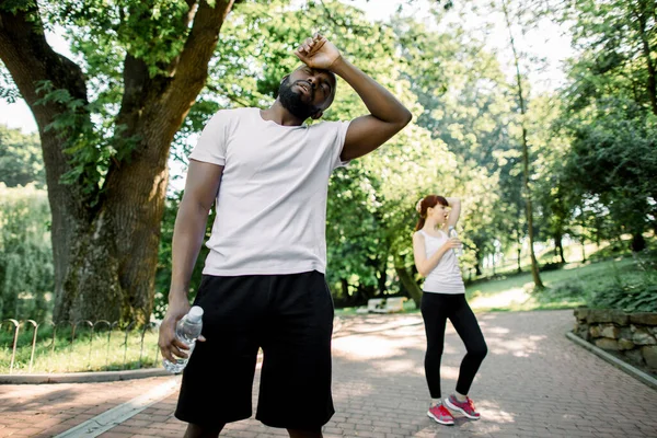 Conceito de estilo de vida desportivo. Homem africano forte jovem cansado após o exercício duro do cardio, estando com a garrafa de água. Garota caucasiana exausta descansando depois de correr no fundo . — Fotografia de Stock