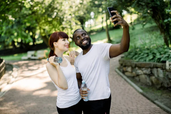 Imagem horizontal de amigos de casal de esportes de fitness multiétnicos no parque ao ar livre, tirando uma foto selfie por telefone celular segurando garrafas com água e sorrindo — Fotografia de Stock