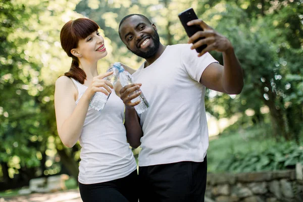 Amigos multiétnicos alegres modernos casal com garrafas de água, fazendo uma pausa para a foto selfie no smartphone, de pé no parque verde urbano durante o treinamento esportivo, jogging ou fitness — Fotografia de Stock