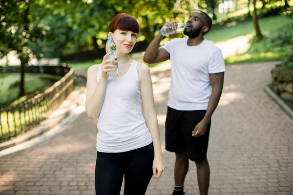 Foto da jovem mulher branca bonita segurando garrafa de água após o treinamento no parque. Bonito homem barbudo africano em pé no fundo no parque de verão enquanto bebe água — Fotografia de Stock