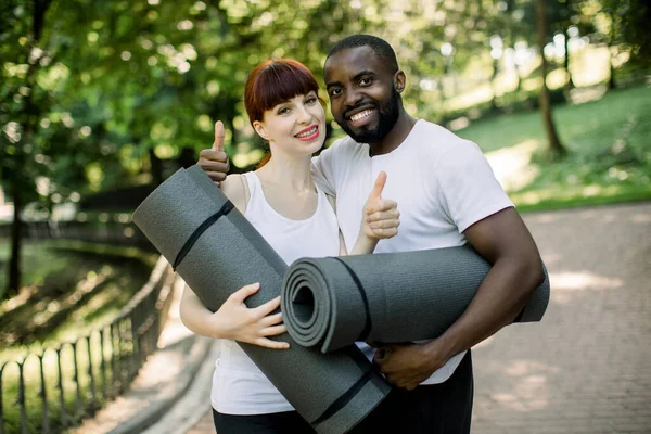 Jovem sorrindo casal multiétnico, menino africano e menina caucasiana, vestindo sportswear, segurando tapetes de ioga, está pronto para se exercitar no parque da cidade ao ar livre, mostrando polegares para cima — Fotografia de Stock