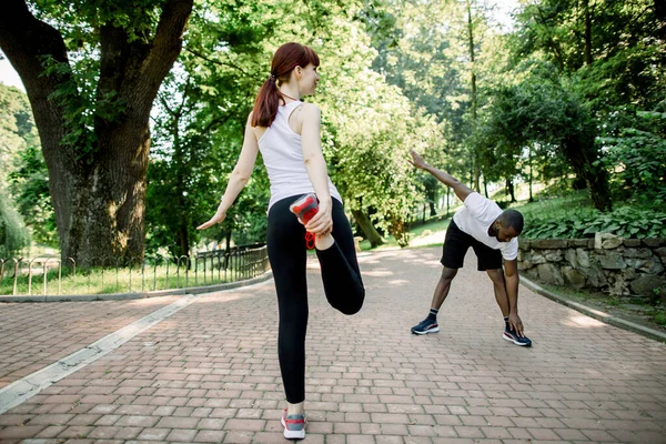 Молодая многонациональная пара бегунов, кавказская рыжая девушка и африканский парень, разогреваются и растягиваются перед утренней пробежкой в городском парке — стоковое фото