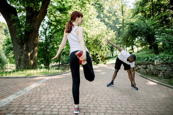 Pasangan pelari multietnis muda, gadis berambut merah kaukasia dan pria Afrika, pemanasan dan peregangan sebelum pagi berjalan di taman kota — Stok Foto