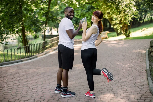 Fitness, sport, friendship and lifestyle concept. Smiling happy multiethnic friends, African boy and Caucasian girl, posing with bottles of water after cardio workout outdoors in city park