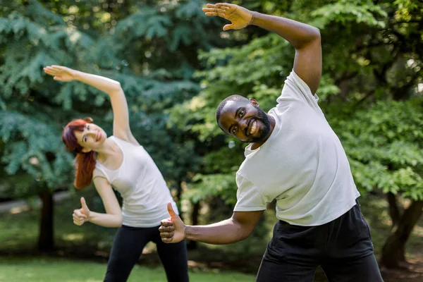 Mulher branca e homem africano, casal fitness, treinando juntos fazendo exercícios corporais de alongamento no parque ao nascer do sol, sorrindo para a câmera e mostrando os polegares para cima. Conceito de deformação desportiva . — Fotografia de Stock