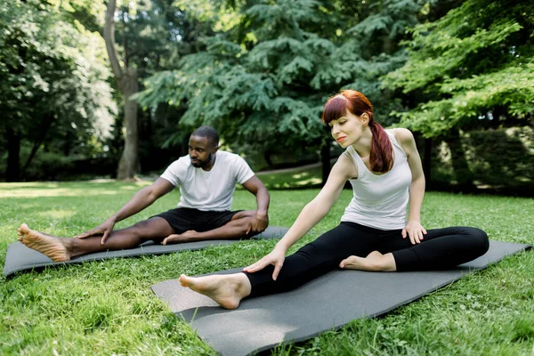 Multiethnic couple doing yoga in nature. Young Caucasian woman performs yoga asanas outdoors in a park, together with her handsome African boyfriend behind. Yoga, fitness, sports, healthy lifestyle