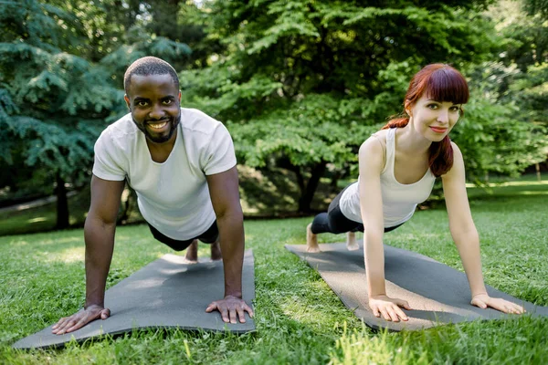 Charming young red haired Caucasian girl and handsome muscular African man, wearing in white t-shirts and black pants doing an exercise plank on mats in a summer green park