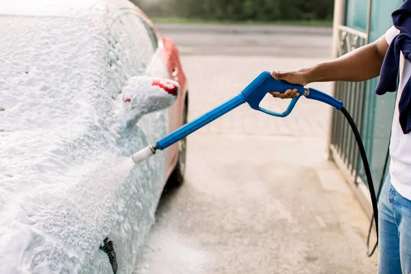 Araba yıkama konsepti. Köpüklü kırmızı araba. Araba sabunla yıkanıyor. Yüksek basınçlı jeti olan Afrikalı bir adam benzin istasyonunda sabunu kırmızı arabaya püskürtüyor. — Stok fotoğraf