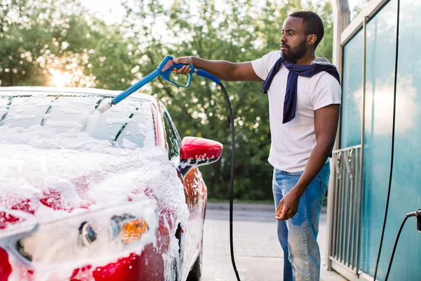 Portrét pohledného vousatého mladého Afričana, jak si myje červené auto pěnou v samoobslužné myčce aut venku. Koncept čistého auta. Myčka aut — Stock fotografie