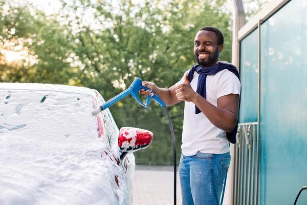 Boldog kacsintó afro-amerikai férfi, a saját autómosó szabadban, permetező tisztító hab egy modern piros luxus autó tartja a magas nyomás és mutatja hüvelykujját felfelé — Stock Fotó