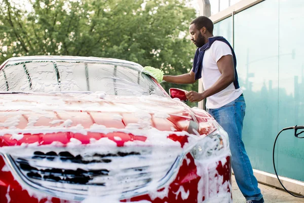 Venkovní mytí aut koncept. Portrét mladého Afričana myjícího si auto v samoobslužné myčce aut s houbou a pěnou — Stock fotografie