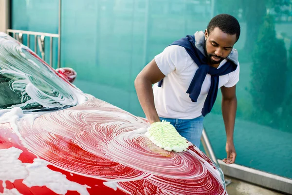 Açık havada oto yıkama servisinin yatay görüntüsü. Sıradan giyinen yakışıklı Afrikalı adam kırmızı arabayı sünger ve köpükle yıkıyor. — Stok fotoğraf