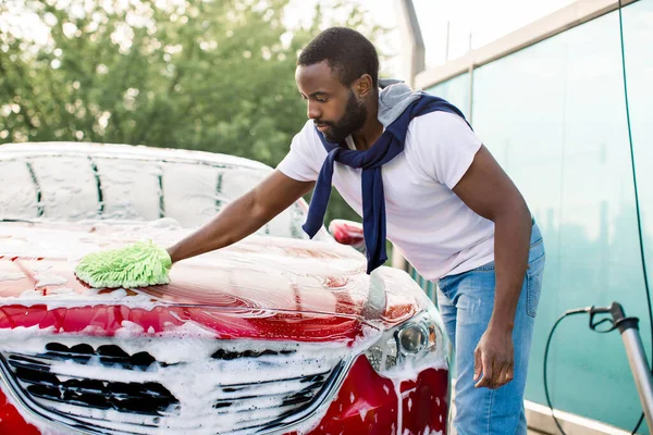 Açık hava oto yıkama servisi. Yakışıklı Afrikalı genç adam kırmızı arabayı yeşil fırça ve köpükle yıkıyor. — Stok fotoğraf