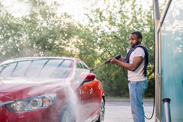 Автомобільна мийка на вулиці. Африканський хлопець у білій футболці та джинсах, прання червоної розкішної машини з водяним пістолетом на автомийці просто неба. — стокове фото