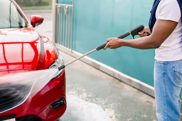Zamknij przycięte poziome zdjęcie rąk Afrykańczyka myjącego swój czerwony luksusowy samochód przy użyciu wody pod wysokim ciśnieniem, na zewnątrz samochodu samoobsługowego mycia — Zdjęcie stockowe