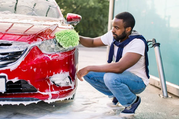 Lavado de coches al aire libre ar estación de servicio de auto lavado. Guapo africano de piel oscura hombre en ropa casual, lavando sus faros rojos con esponja verde y jabón —  Fotos de Stock