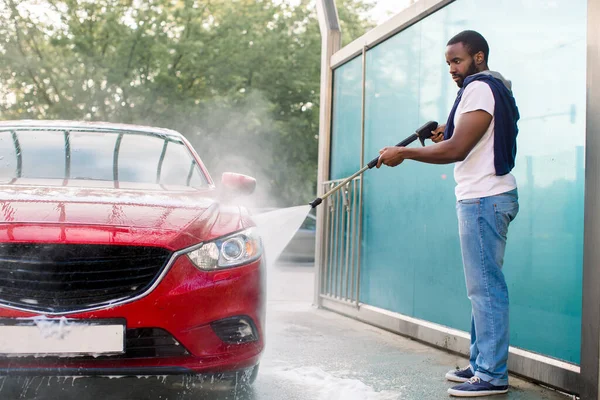Przystojny Afrykanin w t-shircie i dżinsach myjący swój czerwony luksusowy samochód, płukający mydło strumieniem wody pod wysokim ciśnieniem. Ręczna myjnia samochodowa z wodą pod ciśnieniem w myjni samochodowej na zewnątrz. — Zdjęcie stockowe