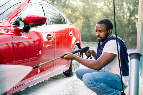 Fiatal afrikai férfi alkalmi viselet mosás a luxus autó saját autómosó állomás szabadban, a nagynyomású víz spray. — Stock Fotó