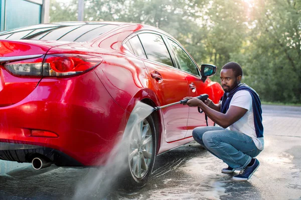 Młody Afrykanin w swobodnym ubraniu myje koła swojego luksusowego samochodu na stacji myjni samoobsługowej na zewnątrz, używając strumienia wody pod wysokim ciśnieniem. Koncepcja myjni samochodowej na zewnątrz — Zdjęcie stockowe