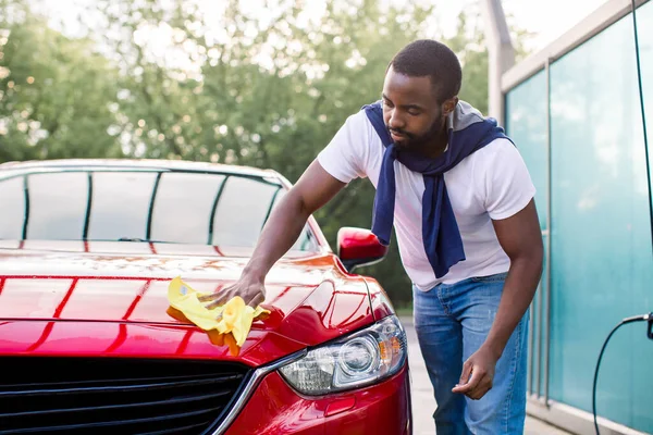 젊은 아프리카계 미국인 남성이 자신의 고급붉은 승용차 실외에서 마이크로 섬유 옷감으로 후드를 청소하는 수평 사진 — 스톡 사진