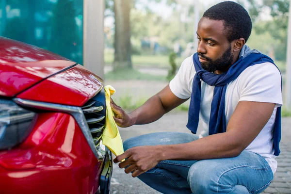 Vista lateral de cerca de la limpieza del coche rojo con paños de microfibra amarilla por un hombre africano barbudo guapo en ropa casual, limpiando la parrilla del radiador del coche en la estación de lavado de automóviles al aire libre —  Fotos de Stock