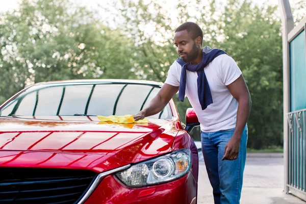 Πλύσιμο αυτοκινήτων και καθαρισμός σε υπαίθριο σταθμό αυτοεξυπηρέτησης. Shot του όμορφος γενειοφόρος νεαρός Αφρικανός καθαρίζοντας το κόκκινο αυτοκίνητο κουκούλα του με ένα κίτρινο πανί μικροϊνών σε εξωτερικούς χώρους το καλοκαίρι ηλιόλουστο πρωί — Φωτογραφία Αρχείου