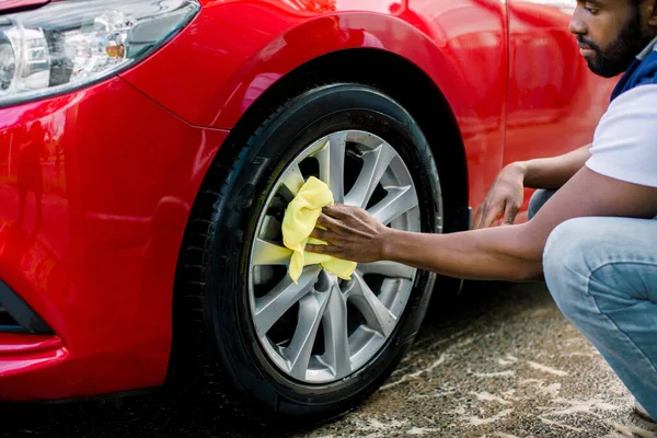 Araba yıkama, fotoğraf çekme, dışarıda araba yıkama. Kırpılmış Afrikalı genç adam görüntüsü, araba yıkamada kırmızı araba alaşımlı tekeri yıkıyor, sarı mikrofiber bez ve özel temizleyici kullanıyor. — Stok fotoğraf