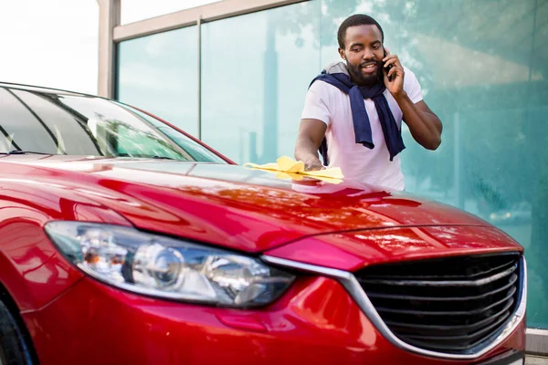 Voiture celaning et polissage à l'extérieur. Portrait de jeune homme africain nettoyant sa voiture avec un chiffon à l'extérieur au lavage de voiture libre-service, et parlant par son smartphone — Photo