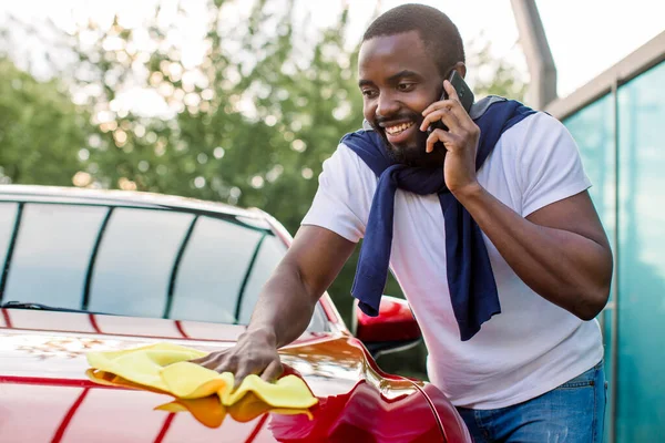 Lavado de coche al aire libre, estación de autoservicio. Joven hombre africano ocupado hablando teléfono mientras pule su coche rojo con tela de microfibra amarilla . — Foto de Stock