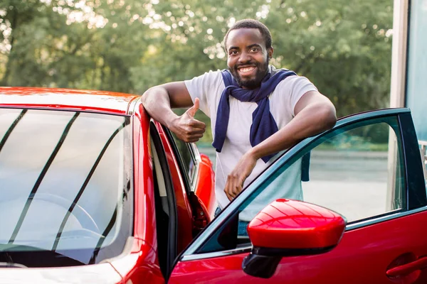 하얀 티셔츠를 입고 스웨터를 입은 아프리카계 미국인 청년이 야외에서 자신의 새 럭셔리 한 붉은 승용차 옆에 서서 엄지손가락을 들고 웃으며 — 스톡 사진