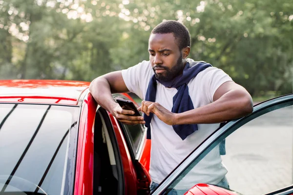 Συγκεντρωμένος νεαρός Αφρικανός που χρησιμοποιεί το έξυπνο τηλέφωνό του για να ελέγξει την κατάσταση ή να ελέγξει την ασφάλεια του αυτοκινήτου του, στέκεται κοντά στο σύγχρονο κόκκινο αυτοκίνητο σε εξωτερικούς χώρους. Εφαρμογές κινητού τηλεφώνου για ιδιοκτήτες αυτοκινήτων. — Φωτογραφία Αρχείου
