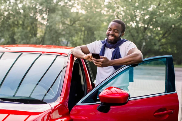 Retrato de estilo de vida al aire libre de la risa satisfecho joven afroamericano en camiseta usando el teléfono móvil, mientras que de pie en su coche rojo moderno. Hombre feliz usando aplicaciones de coche —  Fotos de Stock