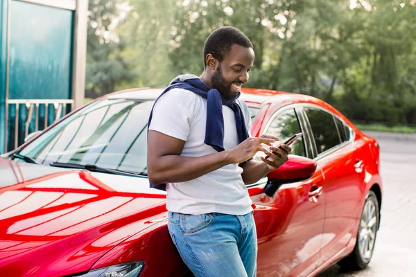 Pria kulit hitam yang santai dan ceria menggunakan telepon pintar dan menulis pesan teks, berdiri di dekat mobil merah modernnya di luar ruangan — Stok Foto
