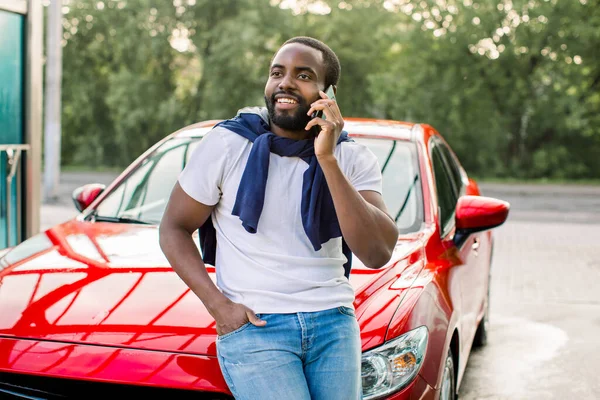 흰색 티셔츠와 청바지를 입은 잘생긴 아프리카 청년이 야외에서 현대의 빨간 자동차 후드를 쓰고 앉아 있는 동안 전화 통화를 하고 있습니다. — 스톡 사진