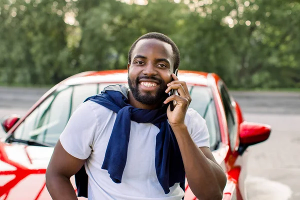 Positive handsome African American man standing in front of his luxury red car and looking at camera with happy smile, while having phone conversation outdoors. Close up horizontal shot