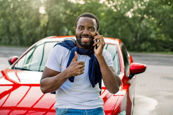 Pemuda Afrika yang gembira dengan pakaian kasual, berdiri di luar ruangan di depan mobil merahnya, berbicara di telepon genggam dan menunjukkan jempolnya ke kamera. Konsep layanan otomatis yang baik, menyalin ruang — Stok Foto