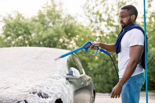Młody czarny człowiek czyści swój szary, luksusowy elektryczny samochód sprayem do piany na zewnątrz w myjni samochodowej. Zwykły Afrykanin myjący samochód opryskiwaczem wysokociśnieniowym i pianką — Zdjęcie stockowe