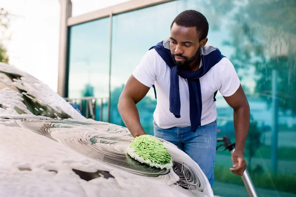 Jonge aantrekkelijke Afro-Amerikaanse man wassen zijn moderne elektrische luxe auto in een self-service car wash station buiten met schoonmaakschuim en groene spons wanten — Stockfoto