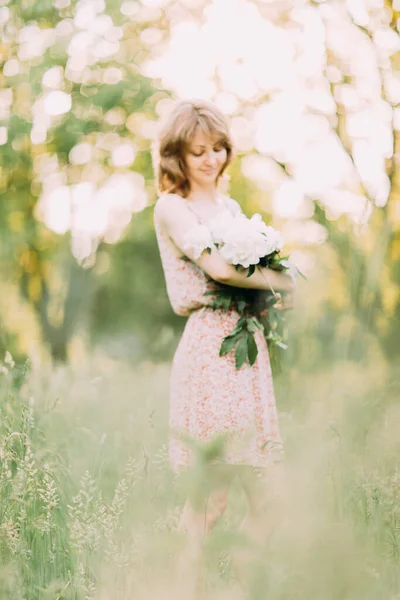 Belle jeune femme blonde caucasienne en robe légère tenant un bouquet de pivoines blanches, se promenant dans un champ d'été ou un jardin au coucher du soleil. Femme avec des fleurs à l'extérieur — Photo