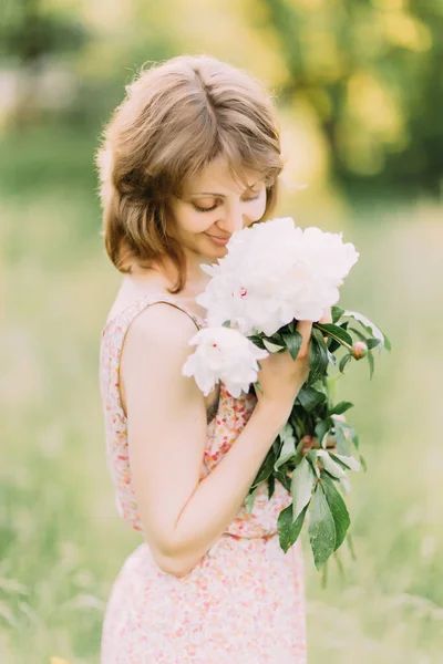 Mulher loira branca bonita em vestido leve segurando buquê de peônias brancas, andando no campo de verão ou jardim ao pôr do sol. Mulher com flores ao ar livre — Fotografia de Stock