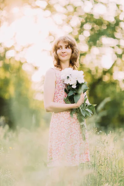 Mulher loira branca bonita em vestido leve segurando buquê de peônias brancas, andando no campo de verão ou jardim ao pôr do sol. Mulher com flores ao ar livre — Fotografia de Stock