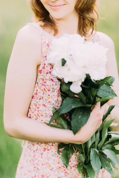 Belle jeune femme blonde caucasienne en robe légère tenant un bouquet de pivoines blanches, se promenant dans un champ d'été ou un jardin au coucher du soleil. Femme avec des fleurs à l'extérieur — Photo