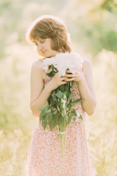 Belle jeune femme blonde caucasienne en robe légère tenant un bouquet de pivoines blanches, se promenant dans un champ d'été ou un jardin au coucher du soleil. Femme avec des fleurs à l'extérieur — Photo