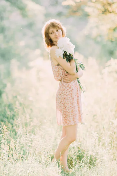 Belle jeune femme blonde caucasienne en robe légère tenant un bouquet de pivoines blanches, se promenant dans un champ d'été ou un jardin au coucher du soleil. Femme avec des fleurs à l'extérieur — Photo
