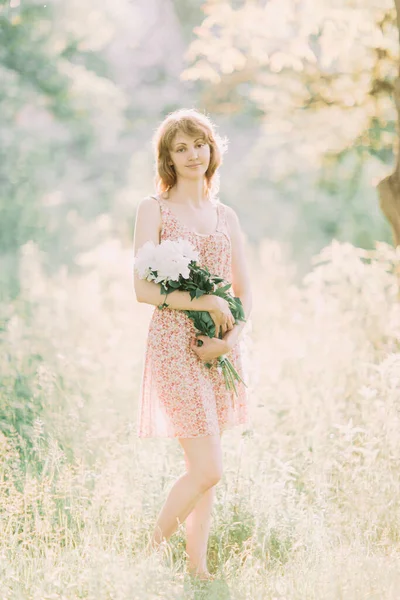 Belle jeune femme blonde caucasienne en robe légère tenant un bouquet de pivoines blanches, se promenant dans un champ d'été ou un jardin au coucher du soleil. Femme avec des fleurs à l'extérieur — Photo