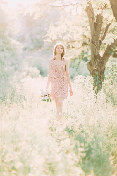 Mulher loira branca bonita em vestido leve segurando buquê de peônias brancas, andando no campo de verão ou jardim ao pôr do sol. Mulher com flores ao ar livre — Fotografia de Stock